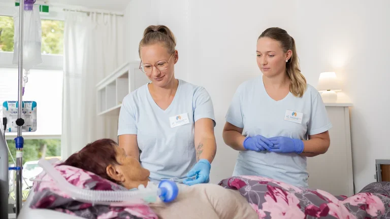 Zwei Pflegerinnen versorgen eine Patientin, die im Bett liegt. 