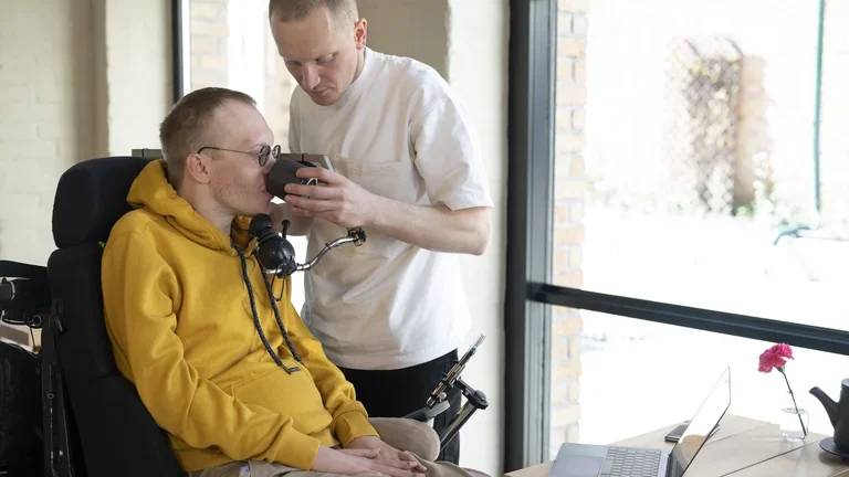 Ein ALS-Patient wird in einer Intensivpflege-WG gepflegt und bekommt von einem Pfleger eine Tasse an seinen Mund geführt.