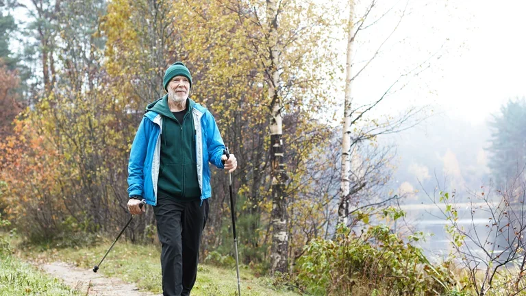 Ein älterer Herr läuft mit Wanderstöcken aktiv und voller Zuversicht einen Waldweg entlang.