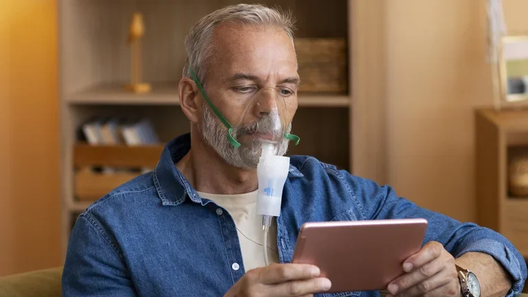 Ein Mann sitzt auf einem Sessel und hat eine Sauerstoffmaske auf. Er hält ein Tablet in der Hand