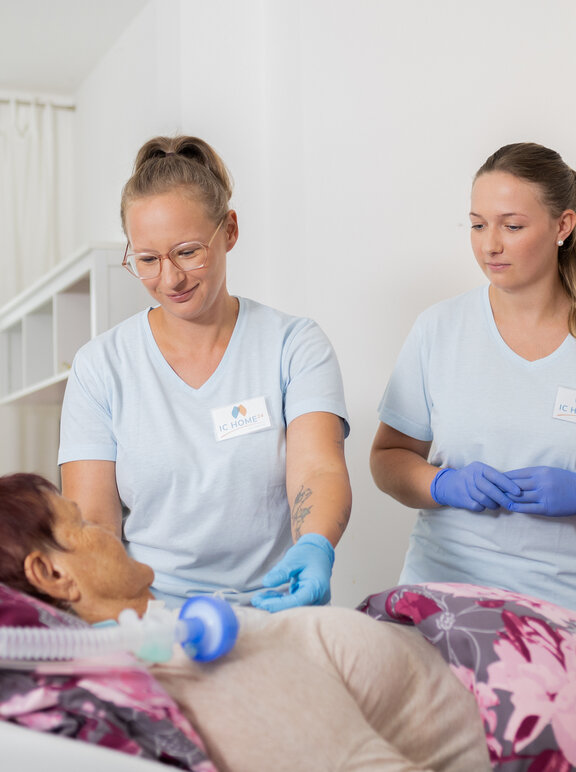 Zwei Pflegerinnen versorgen eine Patientin, die im Bett liegt. 