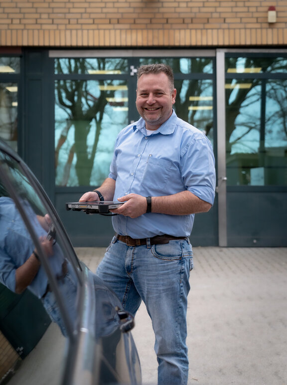 Ein Mann steht mit einem Tablet in der Hand an einem Auto 