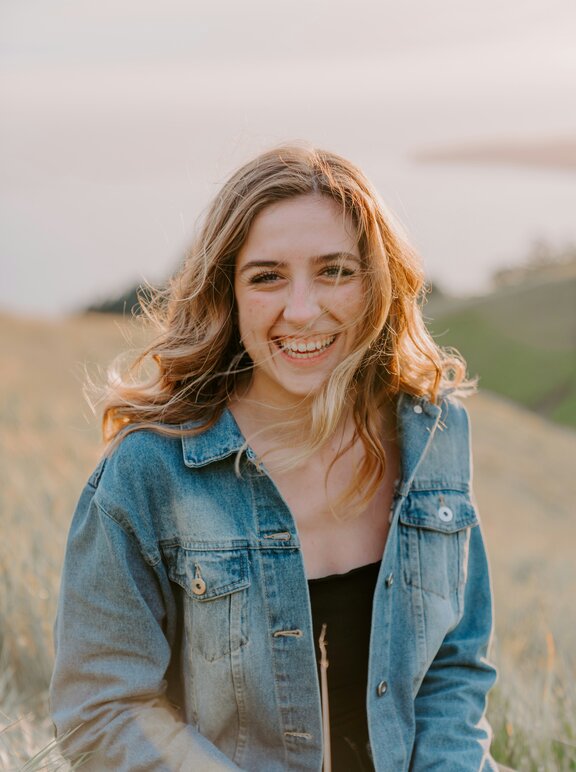 Eine Frau sitzt draußen auf einer Wiese und schaut lachend in die Kamera. 