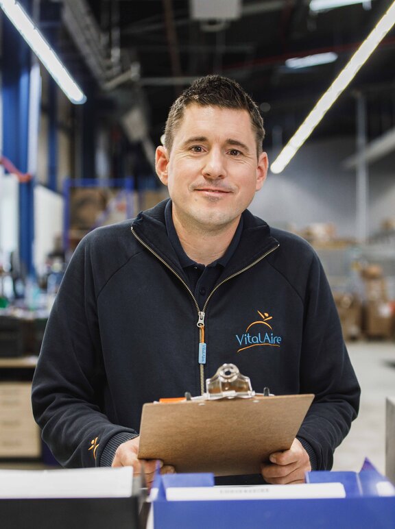 Mitarbeiter steht in einer Lagerhalle und hält ein Klemmbrett in der Hand. Er schaut in die Kamera