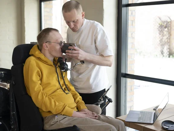 Ein ALS-Patient wird in einer Intensivpflege-WG gepflegt und bekommt von einem Pfleger eine Tasse an seinen Mund geführt.