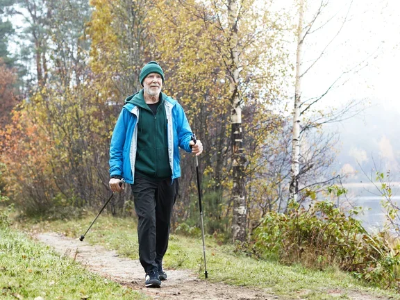 Ein älterer Herr läuft mit Wanderstöcken aktiv und voller Zuversicht einen Waldweg entlang.