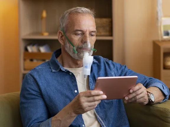 Ein Mann sitzt auf einem Sessel und hat eine Sauerstoffmaske auf. Er hält ein Tablet in der Hand