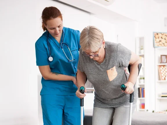 Pflegerin hilft einer Seniorin auf Krücken beim Gehen