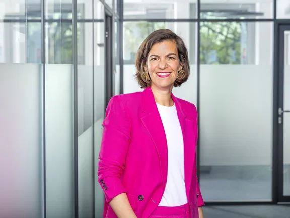 Eine lächelnde Frau im pinken Blazer steht in einem modern gestalteten Büroflur mit Glaswänden.