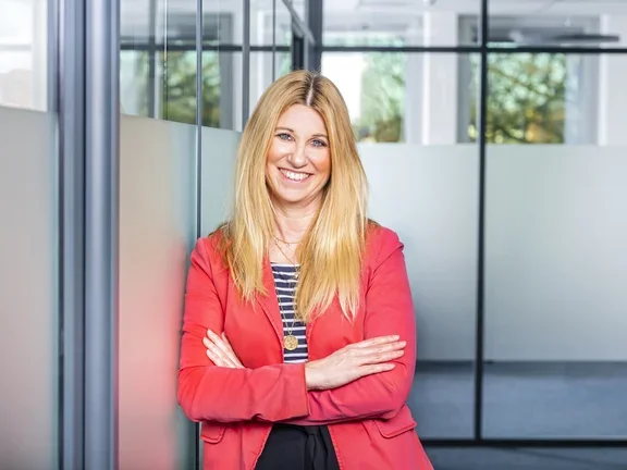 Eine lächelnde Frau im Blazer steht in einem modern gestalteten Büroflur mit Glaswänden.