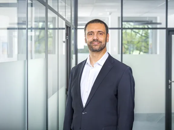 Ein lächelnder Mann im Sakko steht in einem modern gestalteten Büroflur mit Glaswänden.