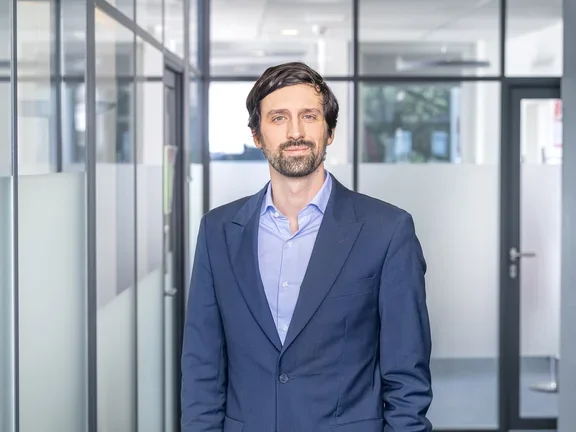 Ein lächelnder Mann im dunkelblauen Sakko und hellblauem Hemd steht in einem modern gestalteten Büroflur mit Glaswänden.