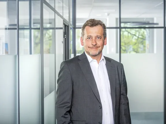 Ein lächelnder Mann im n Sakko steht in einem modern gestalteten Büroflur mit Glaswänden.