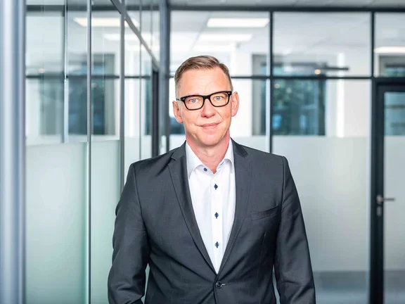 Ein lächelnder Mann im dunkelblauen Sakko steht in einem modern gestalteten Büroflur mit Glaswänden.