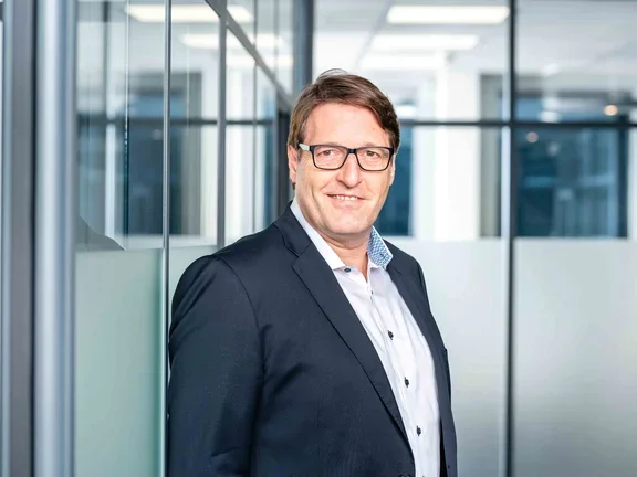 Ein lächelnder Mann im Sakko steht in einem modern gestalteten Büroflur mit Glaswänden.