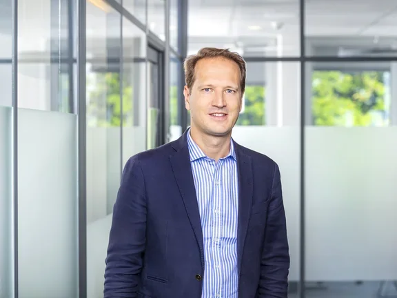 Ein lächelnder Mann im dunkelblauen Sakko und hellblauem Hemd steht in einem modern gestalteten Büroflur mit Glaswänden.