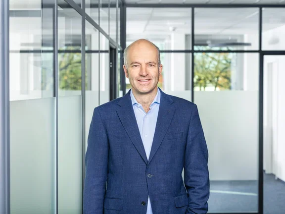 Ein lächelnder Mann im dunkelblauen Sakko steht in einem modern gestalteten Büroflur mit Glaswänden.