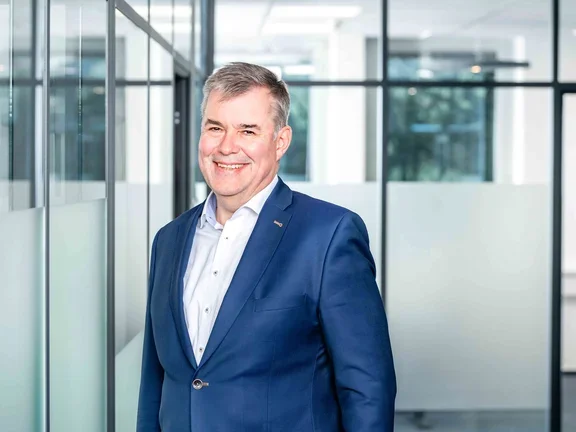 Ein lächelnder Mann im dunkelblauen Sakko und hellblauem Hemd steht in einem modern gestalteten Büroflur mit Glaswänden.