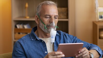 Ein Mann sitzt auf einem Sessel und hat eine Sauerstoffmaske auf. Er hält ein Tablet in der Hand