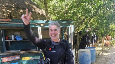 Maik steht in Taucherausrüstung vor der Kamera und hält zwei Finger hoch